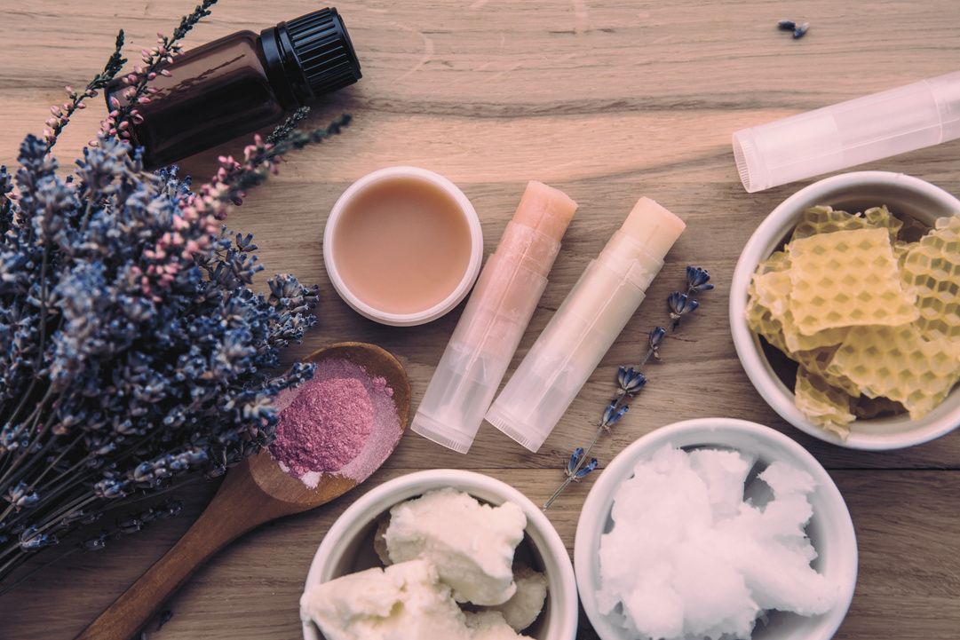 Ingredients for homemade lip balm: shea butter, essential oil, mineral color powder, beeswax, coconut oil. Homemade lip balm lipstick mixture with ingredients scattered around.